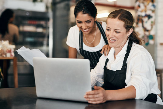 DBS Point of Sale Employee Management Two women using a laptop at a restaurant, showcasing POS system features.